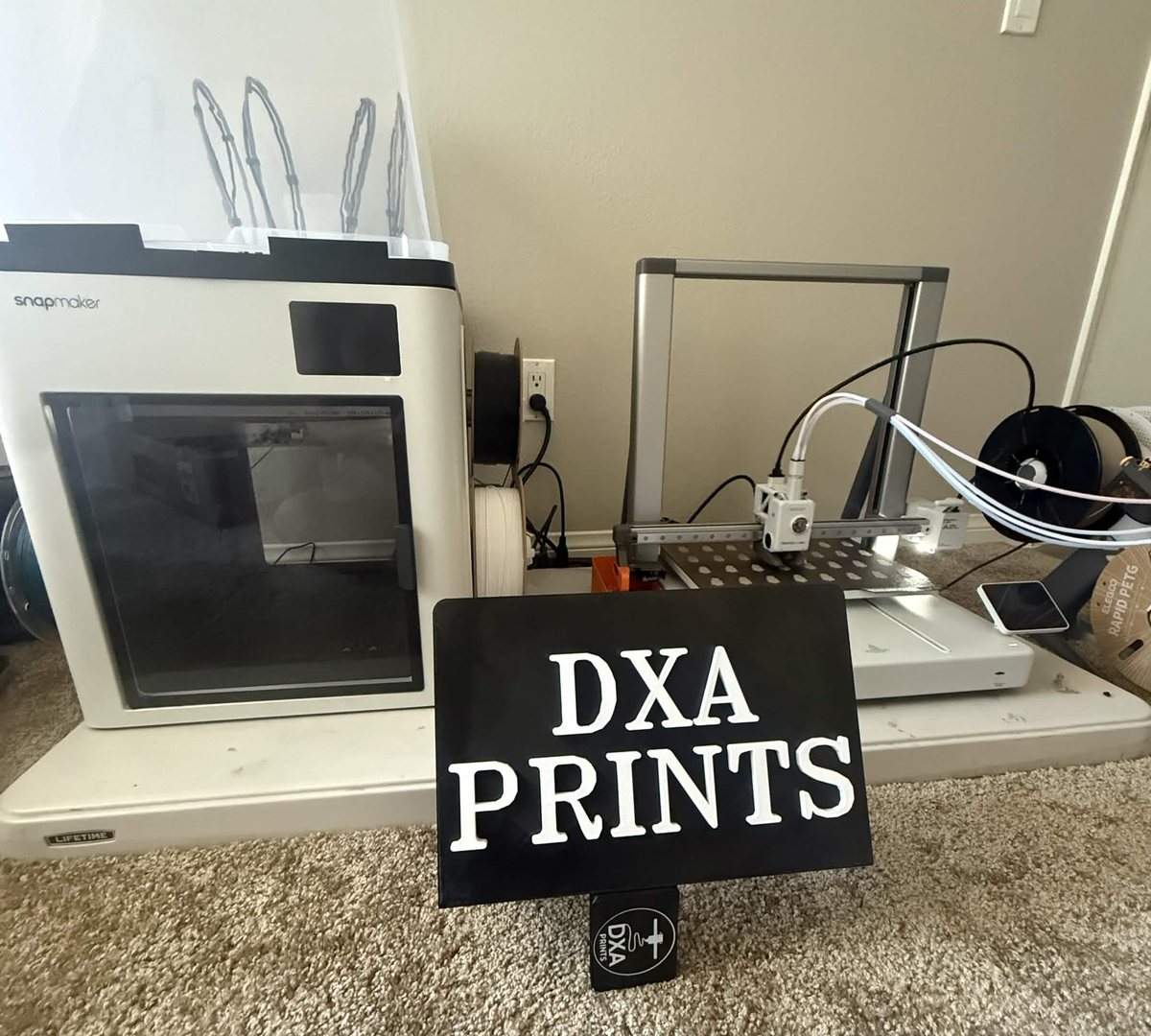 The DXA Prints bench: an enclosed Snapmaker and an open-frame Elegoo printer beside a printed DXA Prints sign.